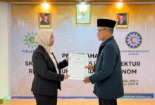 Seorang wanita mengenakan jas hitam dan jilbab menyerahkan dokumen SK resmi kepada seorang pria berjas dan berkopiah hitam di acara PCM Jatinom.
