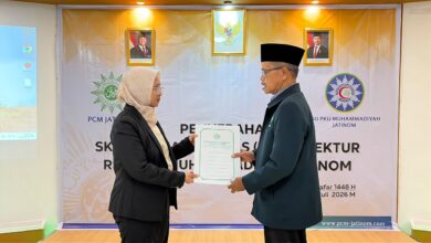 Seorang wanita mengenakan jas hitam dan jilbab menyerahkan dokumen SK resmi kepada seorang pria berjas dan berkopiah hitam di acara PCM Jatinom.