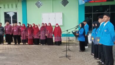 : Foto suasana upacara di halaman kampus yang dihadiri dosen berbatik merah dan mahasiswa berjas almamater biru.