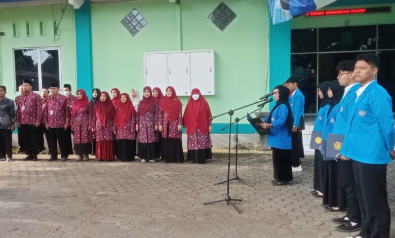 : Foto suasana upacara di halaman kampus yang dihadiri dosen berbatik merah dan mahasiswa berjas almamater biru.