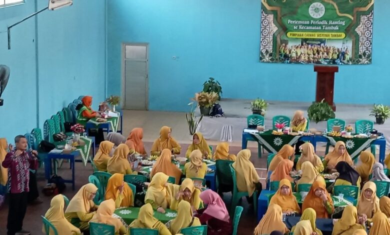 Sekelompok wanita berpakaian kuning dan jilbab senada sedang duduk berkelompok di sekitar meja hijau di dalam ruangan aula yang luas, sedang mengikuti kegiatan pelatihan mitigasi bencana banjir. Di depan, seorang pria berbaju batik memberikan presentasi. Ada spanduk hijau di dinding dengan tulisan "Pertemuan Periodik Ranting se-Kecamatan Tambak". Spanduk tersebut menunjukkan partisipasi 'Aisyiyah dalam kegiatan tersebut. Pemandangan dari atas.