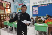 Seorang pria mengenakan pakaian tradisional dan peci hitam berdiri memegang berkas sambil mengajar di depan kelas yang berisi para siswa.