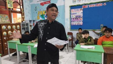 Seorang pria mengenakan pakaian tradisional dan peci hitam berdiri memegang berkas sambil mengajar di depan kelas yang berisi para siswa.