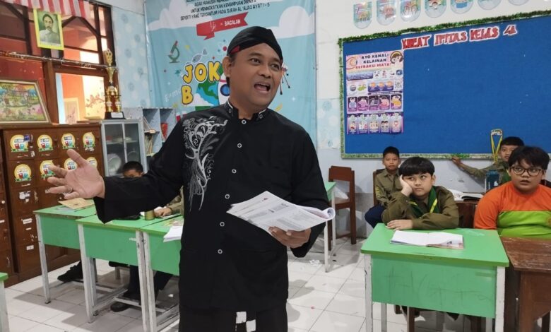 Seorang pria mengenakan pakaian tradisional dan peci hitam berdiri memegang berkas sambil mengajar di depan kelas yang berisi para siswa.