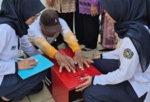 Dua mahasiswi berhijab dari UMS membantu seorang pria paruh baya melakukan tes fleksibilitas menggunakan alat ukur sit and reach berwarna merah.
