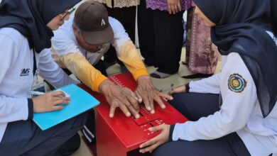 Dua mahasiswi berhijab dari UMS membantu seorang pria paruh baya melakukan tes fleksibilitas menggunakan alat ukur sit and reach berwarna merah.