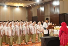 Sekelompok mahasiswa kedokteran berhijab yang baru lulus mengenakan jas lab putih dan rok bermotif batik berdiri dalam barisan menghadap seorang pejabat wanita berhijab merah saat seorang lulusan pria membacakan sumpah dari sebuah buku di auditorium yang terang benderang.