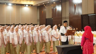 Sekelompok mahasiswa kedokteran berhijab yang baru lulus mengenakan jas lab putih dan rok bermotif batik berdiri dalam barisan menghadap seorang pejabat wanita berhijab merah saat seorang lulusan pria membacakan sumpah dari sebuah buku di auditorium yang terang benderang.