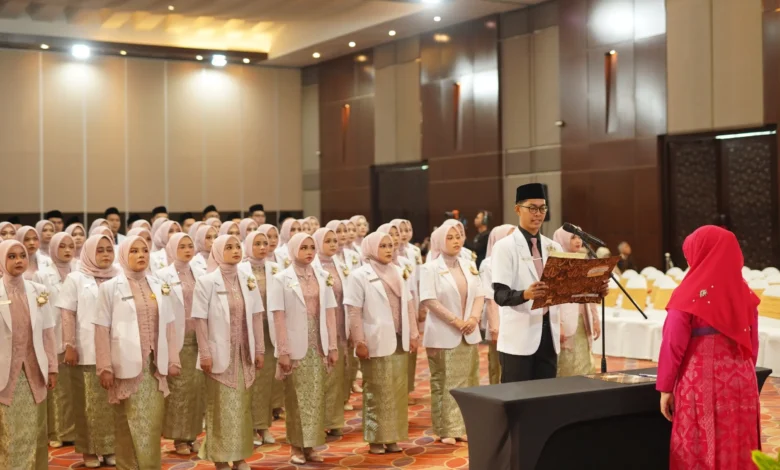 Sekelompok mahasiswa kedokteran berhijab yang baru lulus mengenakan jas lab putih dan rok bermotif batik berdiri dalam barisan menghadap seorang pejabat wanita berhijab merah saat seorang lulusan pria membacakan sumpah dari sebuah buku di auditorium yang terang benderang.