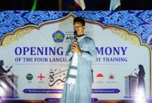 Seorang santri laki-laki di atas panggung besar di malam hari, berbicara dengan percaya diri ke mikrofon. Di belakangnya ada spanduk biru bergaya Arabesque bertuliskan "OPENING CEREMONY OF THE FOUR LANGUAGE ORATORY TRAINING".