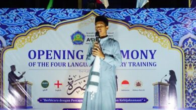 Seorang santri laki-laki di atas panggung besar di malam hari, berbicara dengan percaya diri ke mikrofon. Di belakangnya ada spanduk biru bergaya Arabesque bertuliskan "OPENING CEREMONY OF THE FOUR LANGUAGE ORATORY TRAINING".