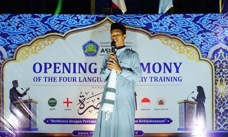 Seorang santri laki-laki di atas panggung besar di malam hari, berbicara dengan percaya diri ke mikrofon. Di belakangnya ada spanduk biru bergaya Arabesque bertuliskan "OPENING CEREMONY OF THE FOUR LANGUAGE ORATORY TRAINING".