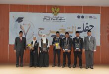 Para wisudawan dan pimpinan institusi berpose bersama di atas panggung dengan latar belakang banner bertuliskan bahasa Arab dan logo wisuda.