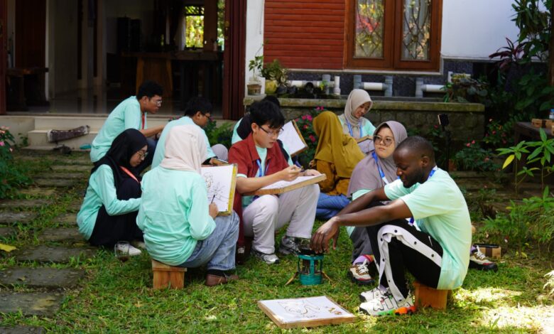 Sejumlah mahasiswa mengenakan kaos seragam hijau toska asyik belajar dan mempraktikkan proses membatik di atas rumput taman bersama peserta asing.