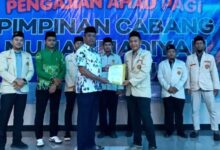 Tangan seorang peserta memegang canting dan menorehkan cairan malam di atas kain putih bermotif batik di dalam ruangan.