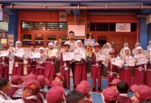Sejumlah siswa berseragam merah putih berdiri berjejer sambil memegang piala, medali, dan sertifikat prestasi di halaman sekolah.