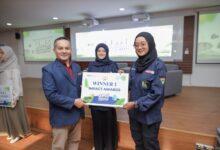 Seorang pria mengenakan jas blazer biru menyerahkan plakat penghargaan bertuliskan Winner 1 Impact Awards kepada mahasiswi berhijab di atas panggung.