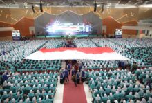 Ribuan mahasiswa baru duduk memenuhi ruang auditorium besar sambil membentangkan bendera merah putih raksasa di bagian tengah.