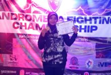 Seorang atlet wanita berhijab mengenakan jaket sport berdiri di depan banner Andromeda Fighting Championship sambil menunjukkan medali dan sertifikat juara.
