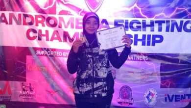Seorang atlet wanita berhijab mengenakan jaket sport berdiri di depan banner Andromeda Fighting Championship sambil menunjukkan medali dan sertifikat juara.