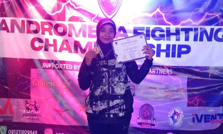 Seorang atlet wanita berhijab mengenakan jaket sport berdiri di depan banner Andromeda Fighting Championship sambil menunjukkan medali dan sertifikat juara.