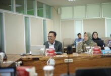 Seorang pria berkacamata mengenakan jas duduk di depan laptop memimpin rapat di ruang sidang bersama rekan kerja.