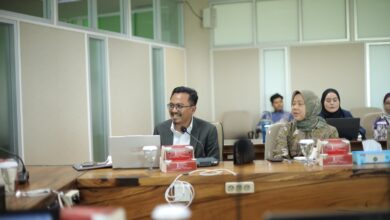 Seorang pria berkacamata mengenakan jas duduk di depan laptop memimpin rapat di ruang sidang bersama rekan kerja.