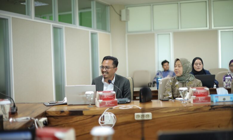 Seorang pria berkacamata mengenakan jas duduk di depan laptop memimpin rapat di ruang sidang bersama rekan kerja.