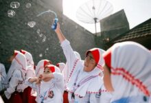Seorang narasumber wanita mengenakan jas almamater berdiri memberikan penyuluhan kesehatan menggunakan proyektor di hadapan para ibu-ibu di dalam ruangan.