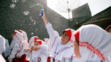 Seorang narasumber wanita mengenakan jas almamater berdiri memberikan penyuluhan kesehatan menggunakan proyektor di hadapan para ibu-ibu di dalam ruangan.