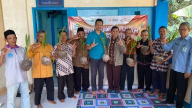 Sembilan pria berpose bersama sambil memamerkan bibit kelapa kopyor yang telah bertunas di depan banner program UMKM.