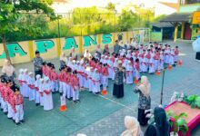 Barisan siswa dan siswi berseragam rapi mengikuti upacara di halaman sekolah berlatar dinding hijau dengan tulisan besar.