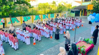 Barisan siswa dan siswi berseragam rapi mengikuti upacara di halaman sekolah berlatar dinding hijau dengan tulisan besar.