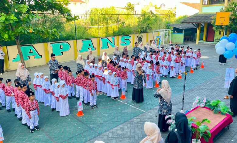 Barisan siswa dan siswi berseragam rapi mengikuti upacara di halaman sekolah berlatar dinding hijau dengan tulisan besar.