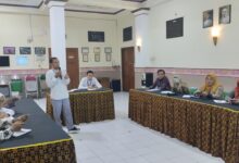 Seorang pria mengenakan kemeja putih memegang mik dan berdiri memberikan pemaparan materi di hadapan peserta rapat di aula.