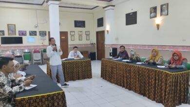 Seorang pria mengenakan kemeja putih memegang mik dan berdiri memberikan pemaparan materi di hadapan peserta rapat di aula.