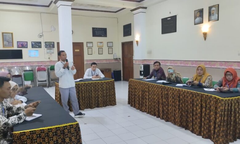 Seorang pria mengenakan kemeja putih memegang mik dan berdiri memberikan pemaparan materi di hadapan peserta rapat di aula.