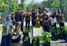 Sekumpulan mahasiswa berpose bersama di dekat monumen patung pohon emas dan polybag bibit tanaman di area taman terbuka.