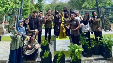 Sekumpulan mahasiswa berpose bersama di dekat monumen patung pohon emas dan polybag bibit tanaman di area taman terbuka.