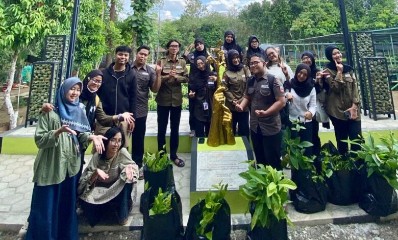 Sekumpulan mahasiswa berpose bersama di dekat monumen patung pohon emas dan polybag bibit tanaman di area taman terbuka.