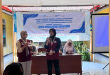 Dua mahasiswi memberikan penyuluhan kesehatan di depan banner PPK Ormawa HMPSIK Universitas Muhammadiyah Purwokerto di balai desa.