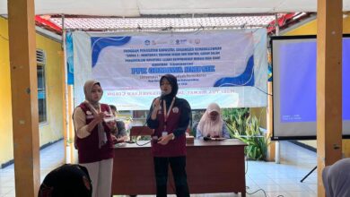 Dua mahasiswi memberikan penyuluhan kesehatan di depan banner PPK Ormawa HMPSIK Universitas Muhammadiyah Purwokerto di balai desa.