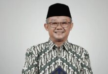 Portrait Mendikdasmen Abdul Mu'ti mengenakan kemeja batik dan peci hitam tersenyum memandang kamera dengan latar belakang polos.
