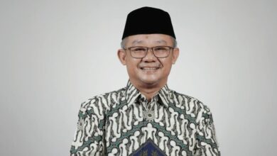 Portrait Mendikdasmen Abdul Mu'ti mengenakan kemeja batik dan peci hitam tersenyum memandang kamera dengan latar belakang polos.