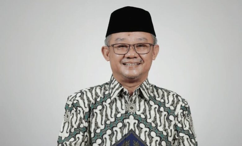 Portrait Mendikdasmen Abdul Mu'ti mengenakan kemeja batik dan peci hitam tersenyum memandang kamera dengan latar belakang polos.