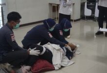 Tiga orang mahasiswa berkerudung biru, berjaket PMI, dan bermasker, sedang memberikan pertolongan pertama kepada seseorang yang berbaring di lantai, disaksikan oleh juri.