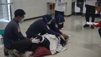 Tiga orang mahasiswa berkerudung biru, berjaket PMI, dan bermasker, sedang memberikan pertolongan pertama kepada seseorang yang berbaring di lantai, disaksikan oleh juri.