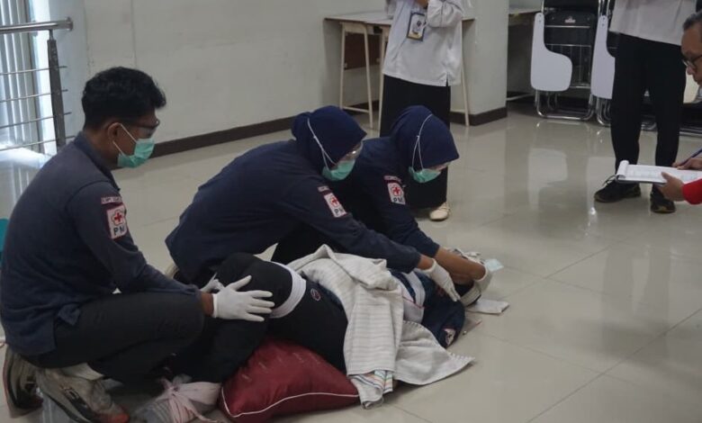 Tiga orang mahasiswa berkerudung biru, berjaket PMI, dan bermasker, sedang memberikan pertolongan pertama kepada seseorang yang berbaring di lantai, disaksikan oleh juri.