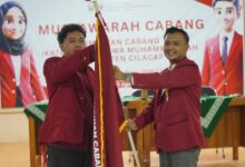 Dua pria berjas almamater merah sedang melakukan prosesi serah terima bendera pataka IMM dalam acara Musyawarah Cabang IMM Cilacap.