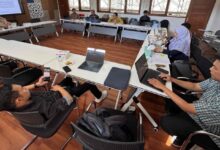 Suasana rapat revisi kurikulum PTI UMS bersama dosen dan staf di ruang rapat dengan lantai kayu. Tampak layar monitor menampilkan dokumen akademik dan beberapa peserta menggunakan laptop serta smartphone.